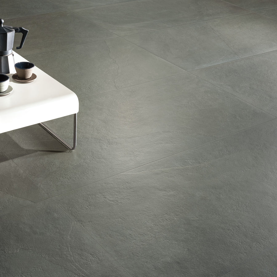 A minimalist room with large gray stone tiles on the floor. A white table with metal legs holds a coffee maker, cups, and a bowl in the upper left corner of the image.