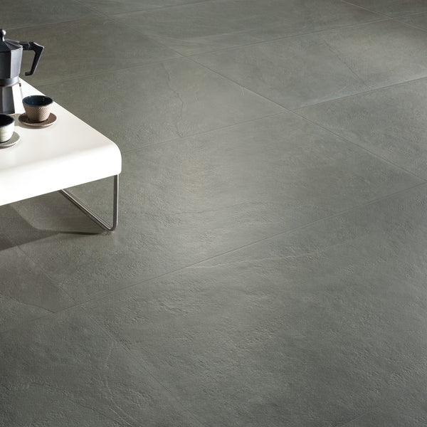 A minimalist room with large gray stone tiles on the floor. A white table with metal legs holds a coffee maker, cups, and a bowl in the upper left corner of the image.