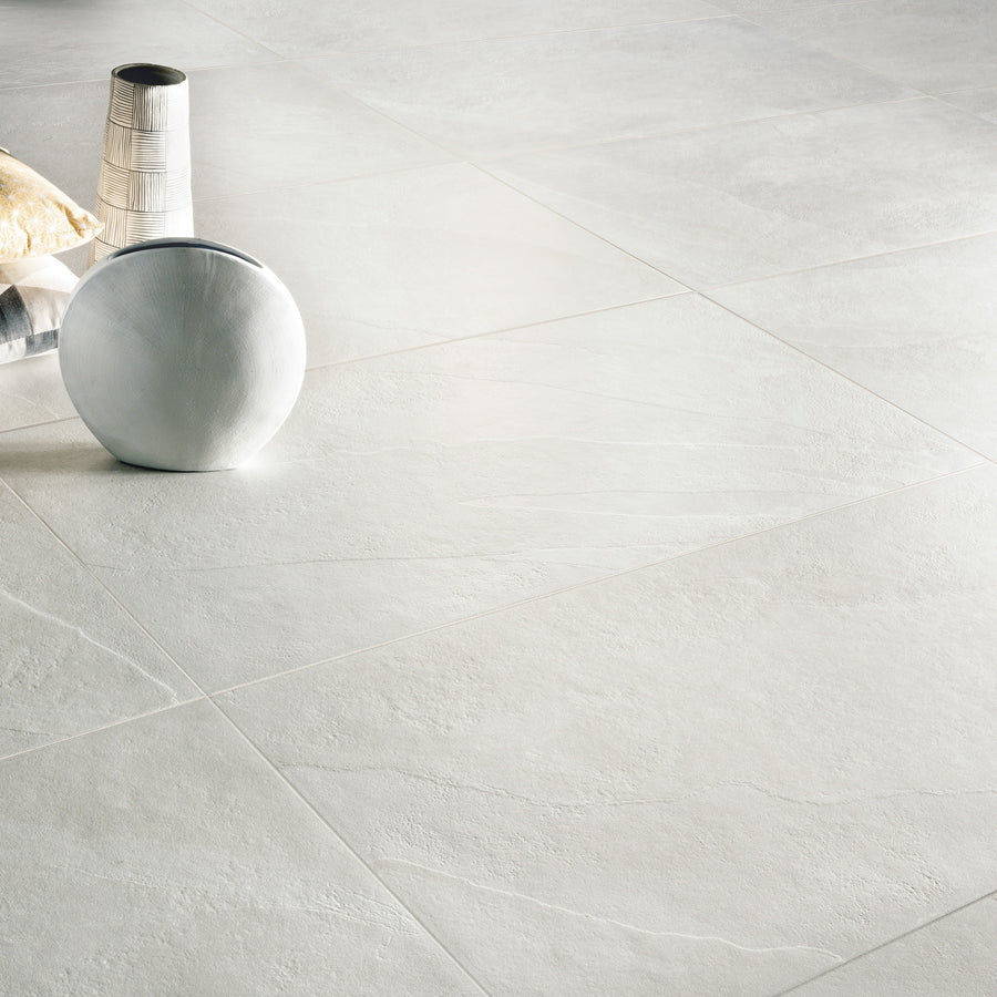 Large light gray stone floor tiles with subtle texture. Decorative items, including a striped vase, a round white cushion, and a beige pillow, are placed on the floor in the top left corner of the image.