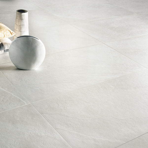 Large light gray stone floor tiles with subtle texture. Decorative items, including a striped vase, a round white cushion, and a beige pillow, are placed on the floor in the top left corner of the image.