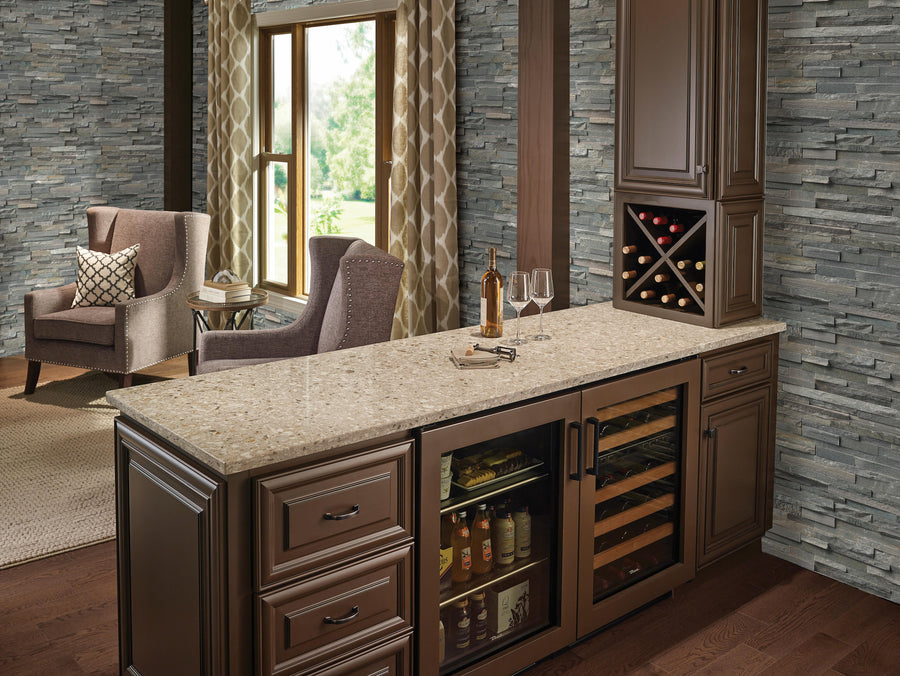 A modern home bar with brown cabinets, a granite countertop, a wine rack, mini fridges with drinks, and two wine glasses. Two armchairs and large windows are in the background, set against gray stone walls.