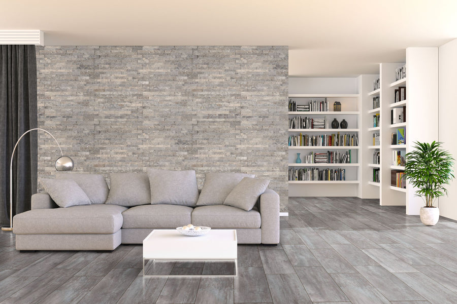 Modern living room with a gray sectional sofa, floor lamp, white coffee table, stone accent wall, built-in bookshelves filled with books, and a green potted plant on gray tiled floors.