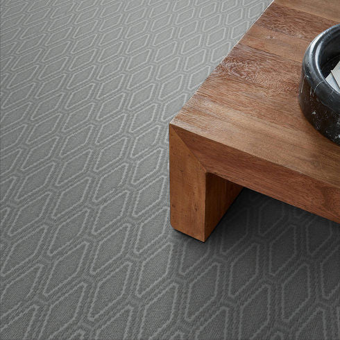 A close-up view of a gray carpet with a geometric diamond pattern, next to a corner of a wooden table with a black round object placed on top.