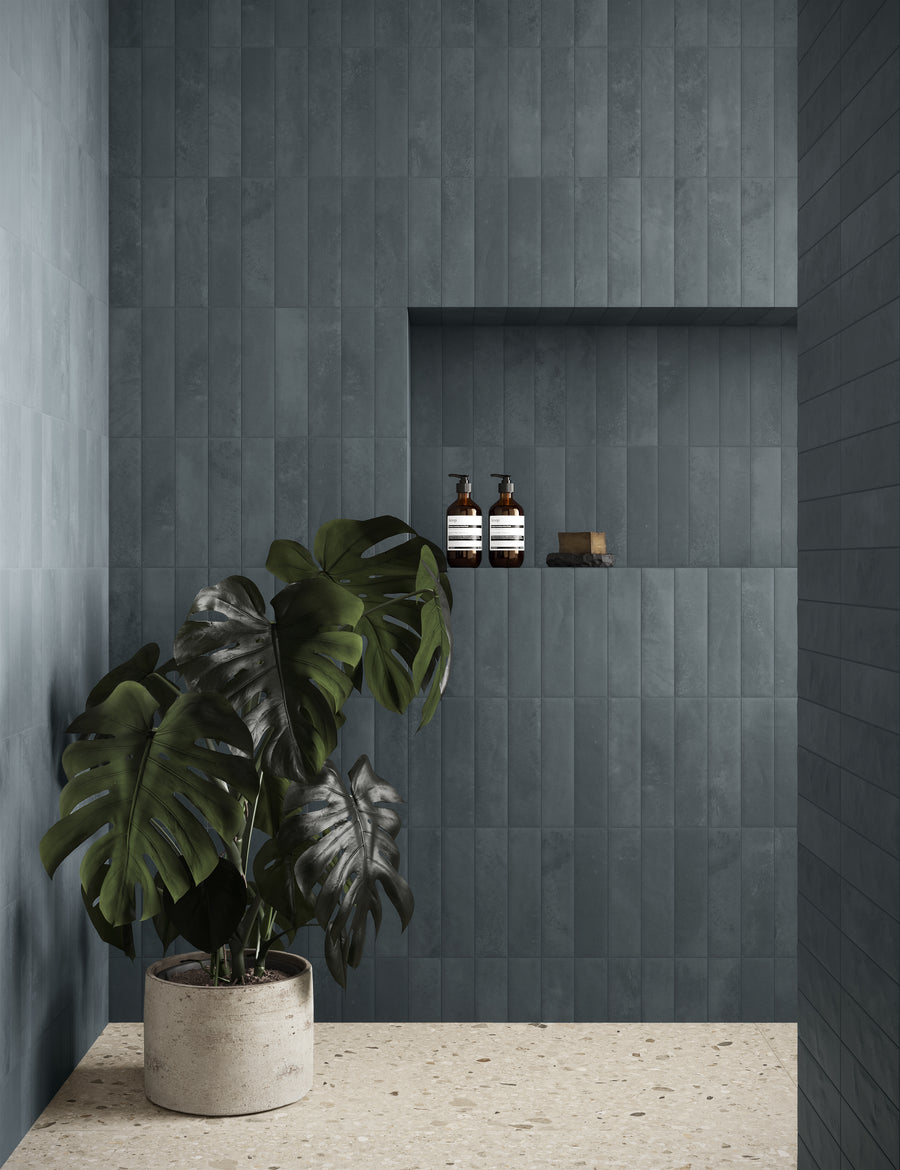 A modern bathroom with matte blue-grey rectangular tiles, a terrazzo floor, a large potted monstera plant, and a recessed wall shelf holding bottles and a sponge.
