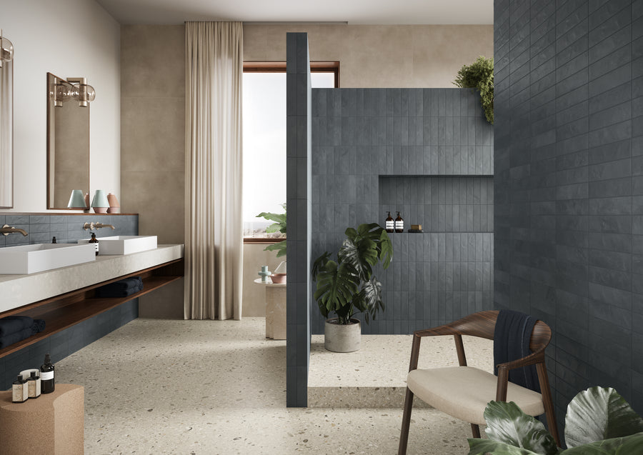 Modern bathroom with blue-grey tiled shower area, double sinks with large mirrors, a wooden chair with a dark throw, terrazzo flooring, potted plants, and beige curtains over a window letting in natural light.