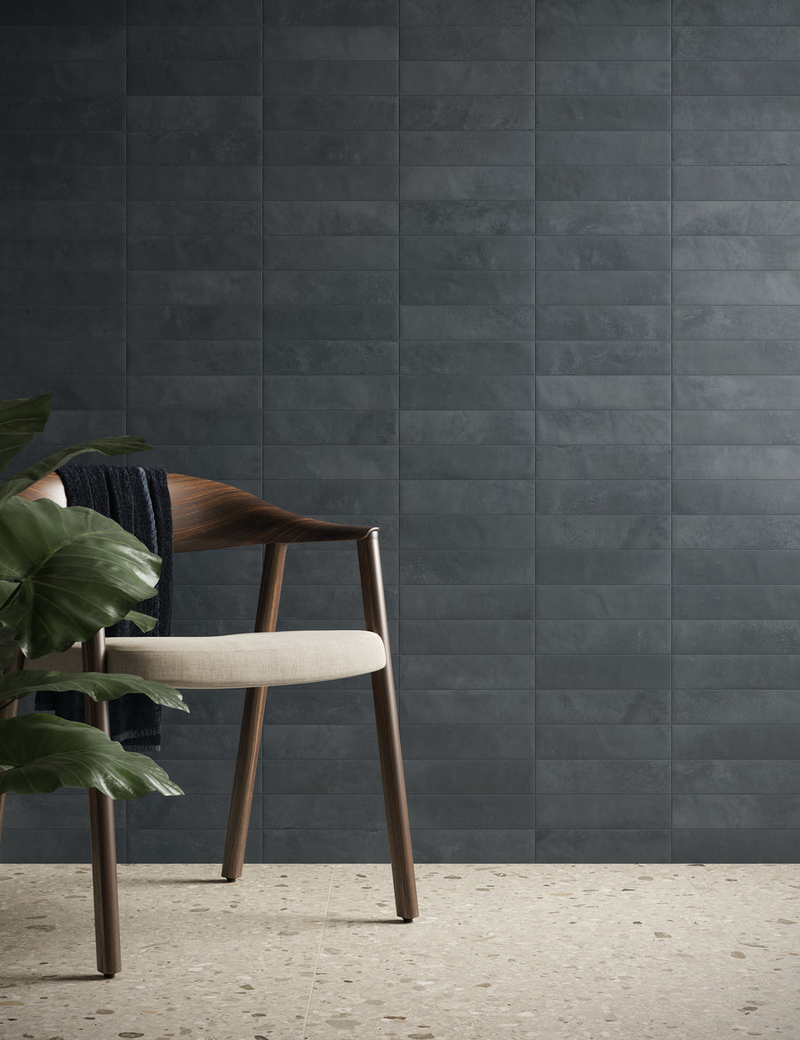 A modern wooden chair with a light cushioned seat sits next to green leafy plants against a dark, textured tiled wall and a light speckled floor. A dark fabric is draped over the chairs armrest.