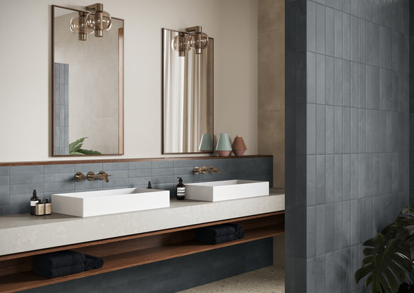 Modern bathroom with two rectangular white sinks, gold faucets, and twin mirrors above a countertop. Decor includes bottles, candles, and folded towels on an open wooden shelf beneath the sinks. Blue tiled backsplash and neutral walls.
