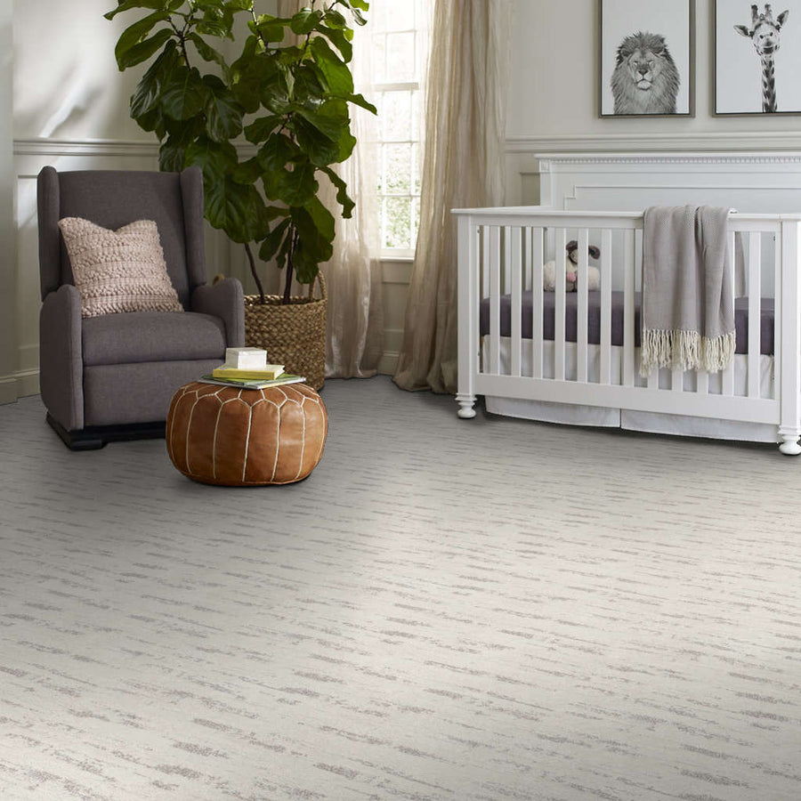 A cozy nursery with a gray armchair, a brown leather pouf, a white crib with a gray blanket, wall art with animal illustrations, and a large leafy plant near a window.