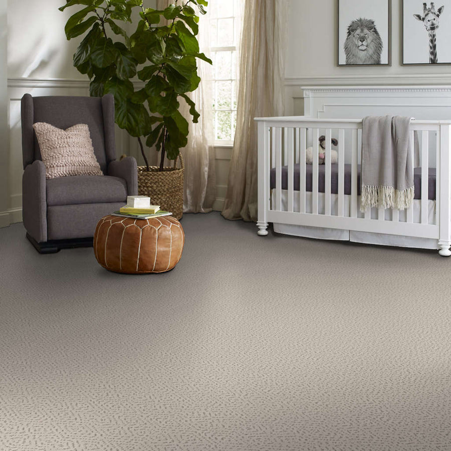 A cozy nursery with a gray armchair, a brown leather pouf, a crib with a gray blanket, a large potted plant, and animal art on the wall. Sunlight streams in through a window with white curtains.
