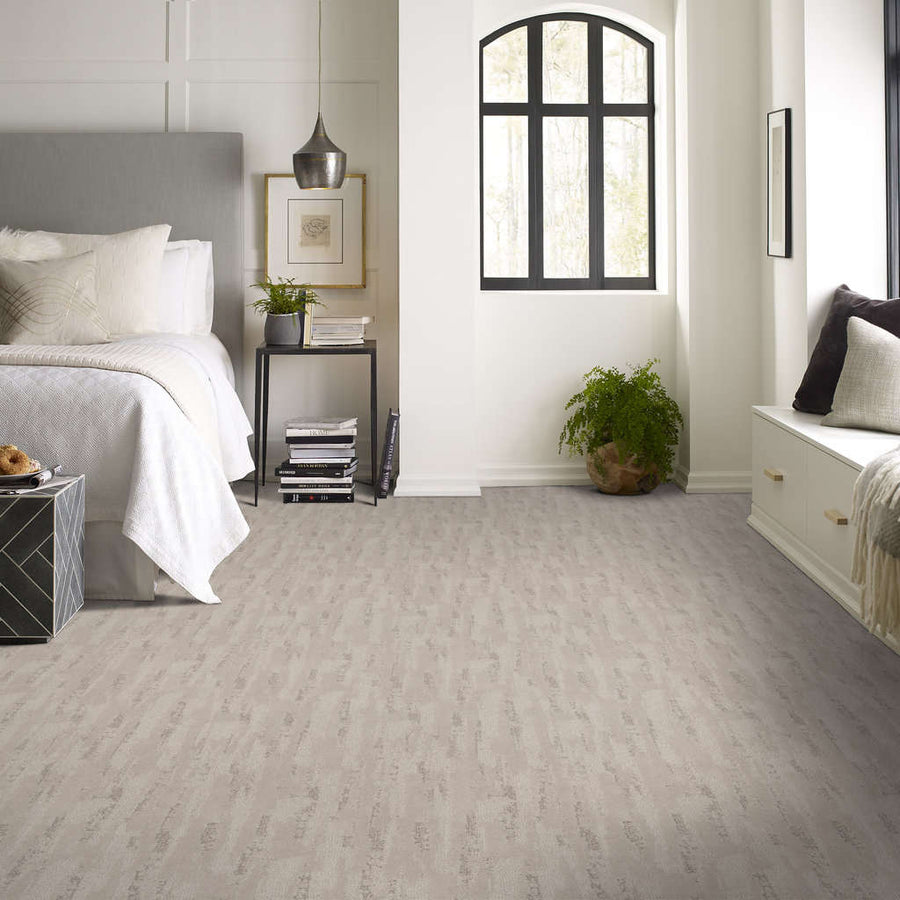 Modern bedroom with a soft gray carpet, white walls, a gray upholstered bed, minimal decor, black side table, green plant, and large black-framed window letting in natural light.
