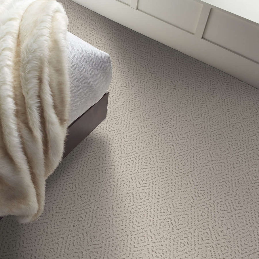 Close-up of a bedroom corner with a bed covered in a textured white blanket and a beige carpet featuring a subtle geometric pattern. Sunlight is coming in from a nearby window.