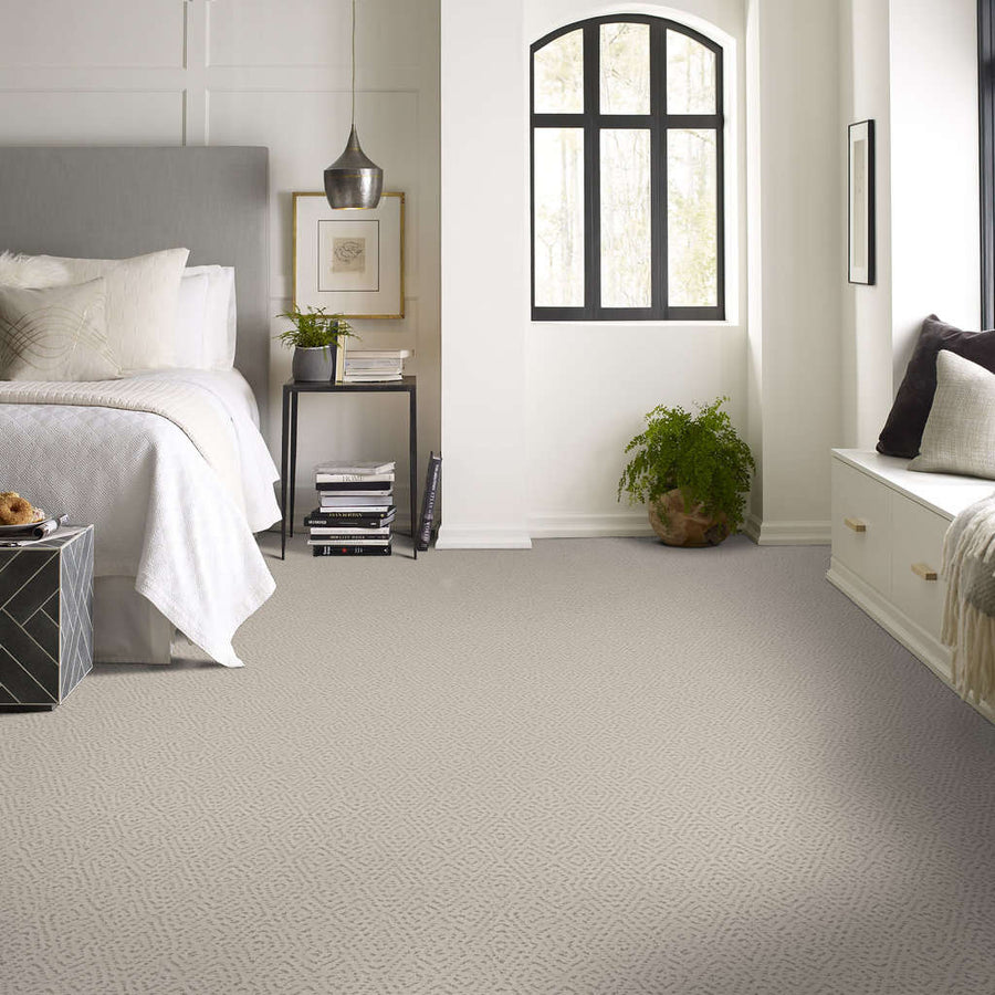 A bright, modern bedroom with light gray carpet, a neatly made bed with white linens, a black bedside table with a lamp and decor, a potted plant, a window seat, and large arched windows letting in natural light.