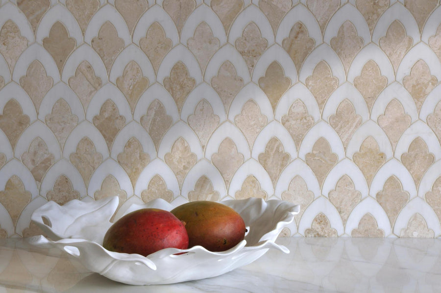 A white decorative bowl holding two mangos sits on a marble surface in front of a beige and white patterned tile backsplash.
