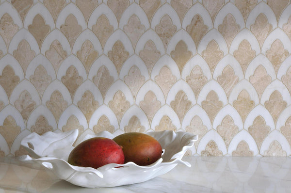 A white decorative bowl holding two mangos sits on a marble surface in front of a beige and white patterned tile backsplash.