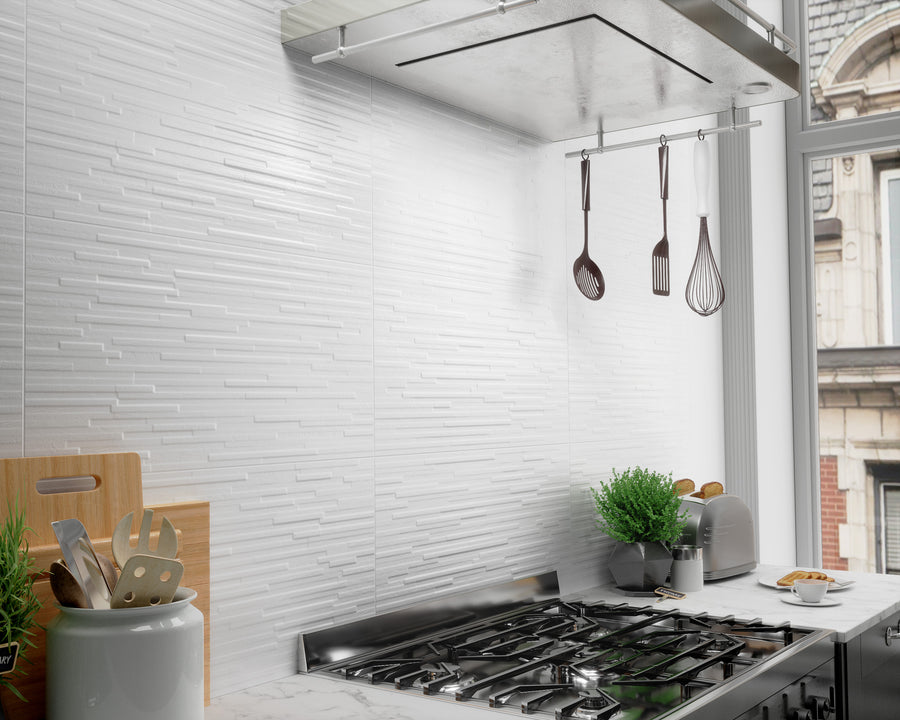 A modern kitchen with a stainless steel gas stove, hanging utensils, potted plants, and a white textured tile backsplash. Light streams in from a large window beside the countertop.