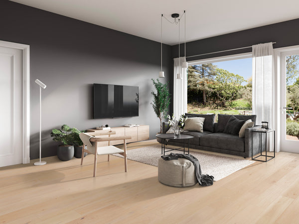 Modern living room with light wood floor, gray accent wall, dark sofa, round coffee table, white rug, potted plants, TV, and large window with sheer curtains overlooking a green outdoor view.
