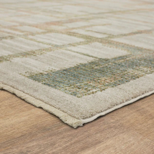 A close-up of a rectangular area rug with a geometric pattern in muted tones of beige, green, and blue, lying on a light wood floor. The rug’s corner and textured surface are clearly visible.