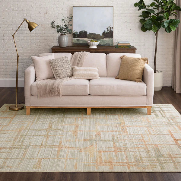 A light beige sofa with throw pillows and a blanket sits on a patterned rug. Behind it, there’s a wooden console table with decor and a painting, a floor lamp on the left, and a tall potted plant on the right against a white brick wall.