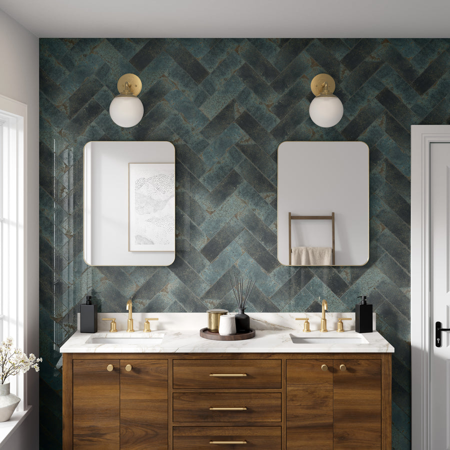 Modern bathroom with a double sink vanity, wood cabinets, gold fixtures, two rectangular mirrors, globe wall lights, and a blue-green herringbone tile accent wall. A towel rack and window are also visible.