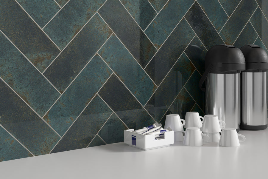 A kitchen counter with stacked white mugs, two stainless steel airpot coffee dispensers, and a box of individual sugar packets, set against a dark teal herringbone tile backsplash.