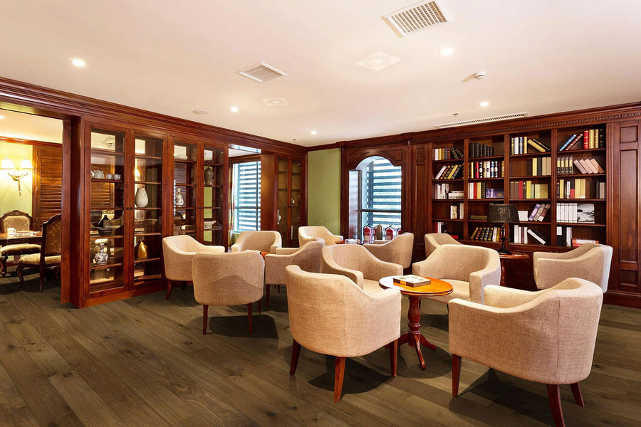 A cozy library lounge with wooden bookshelves, glass cabinets, cream-colored armchairs arranged around small tables, and large windows letting in natural light.