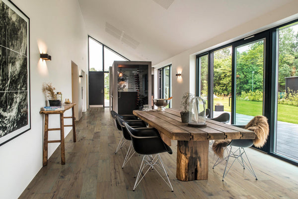 A modern dining room with a rustic wooden table, black chairs, large windows overlooking a green yard, wooden floors, white walls, and minimalist decor including a wall art piece and small table with books.