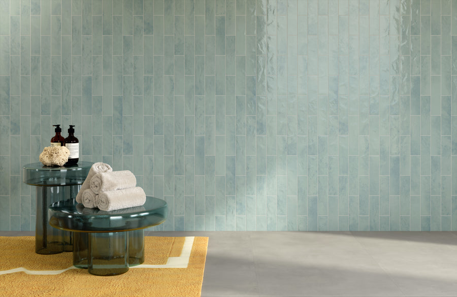 Two round glass tables with rolled white towels, a sponge, and toiletries sit on a yellow rug and gray floor, in front of a wall covered with glossy, light blue vertical tiles.