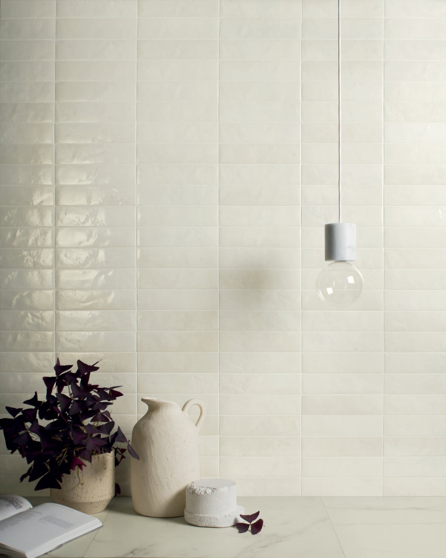 A minimalist scene featuring a glossy cream tile wall, a white marble countertop with a ceramic pitcher, a round container, a book, a potted purple-leafed plant, and a hanging light bulb with a white fixture.