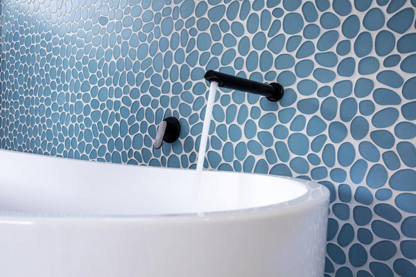 Close-up of a modern bathtub with water flowing from a black wall-mounted faucet. The background features blue and white pebble-shaped mosaic tiles.