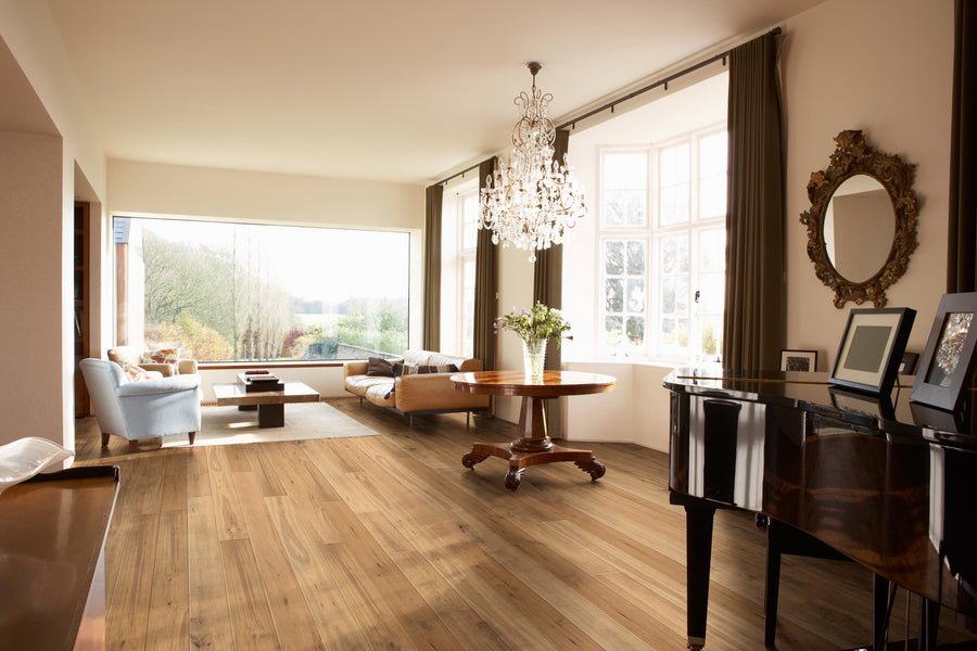A spacious, elegant living room with wooden floors, a grand piano, chandelier, round table with flowers, armchairs, sofa, large windows, and abundant natural light. A gold-framed mirror hangs on the wall.