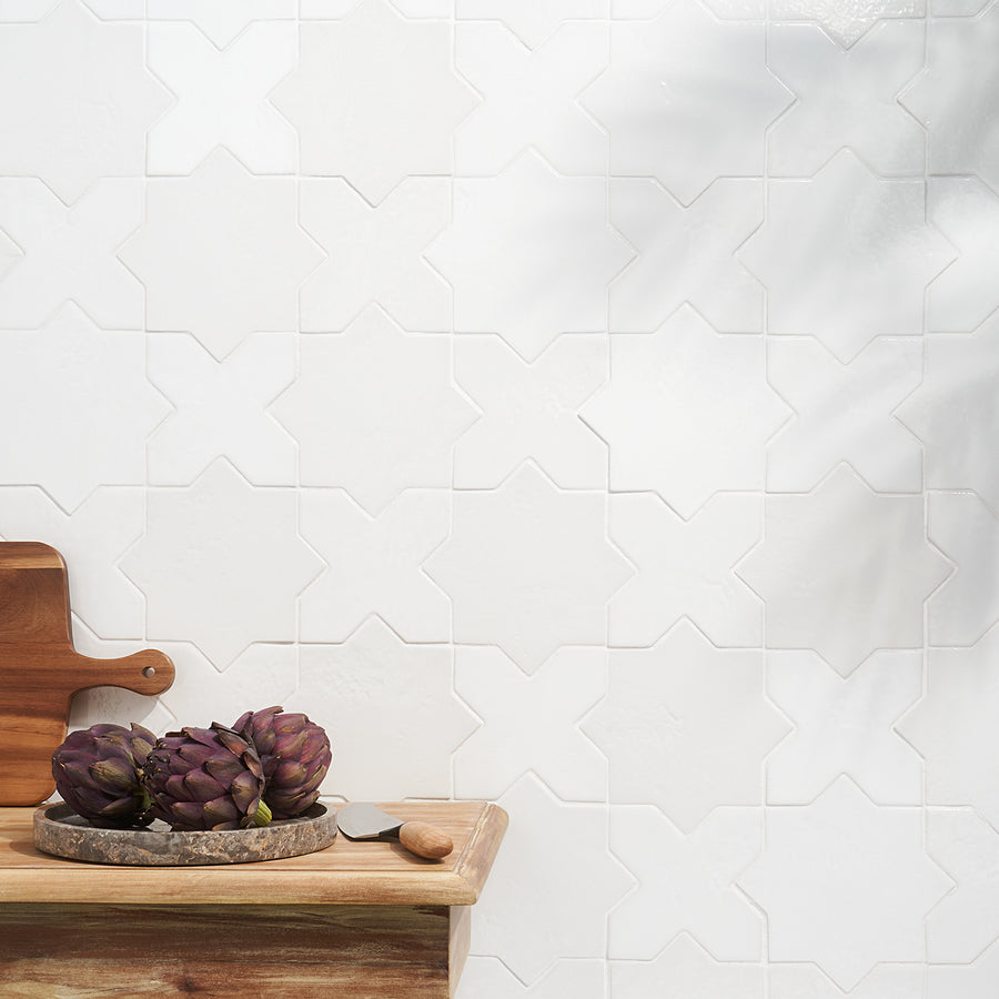 A wooden table with a cutting board, a knife, and three artichokes sits in front of a white, geometric-patterned tiled wall. Soft light and shadows fall across the scene.