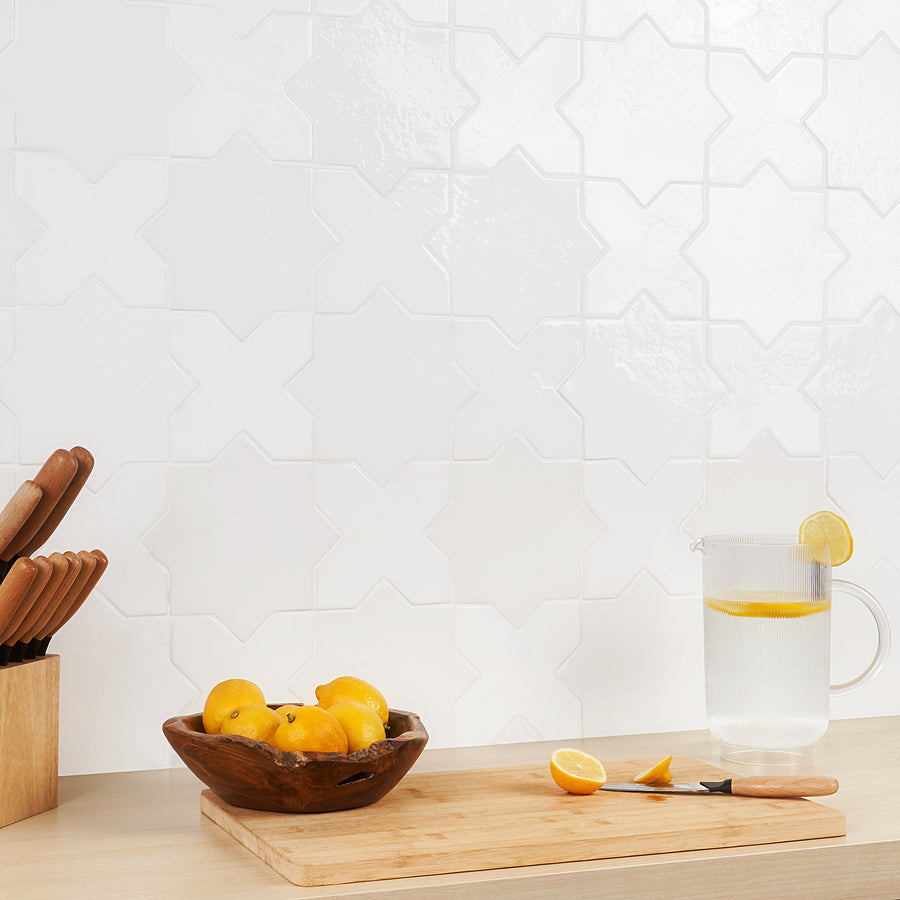 A wooden bowl of lemons, a knife, and a glass pitcher of lemon water sit on a light wood countertop against a white, textured geometric tile backsplash. A wooden knife block is on the left.