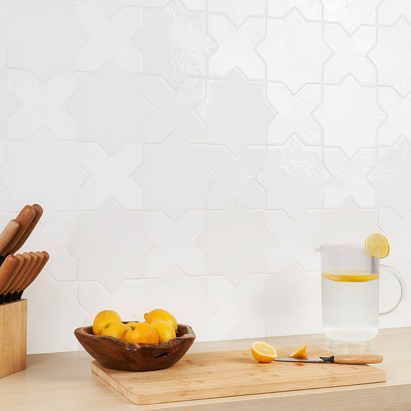 A wooden bowl of lemons, a knife, and a glass pitcher of lemon water sit on a light wood countertop against a white, textured geometric tile backsplash. A wooden knife block is on the left.