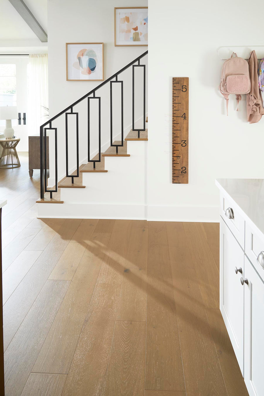 A modern, bright entryway with light wood floors, white walls, a black metal staircase railing, framed abstract art, a large wooden growth chart, and a pink backpack hanging on the wall.