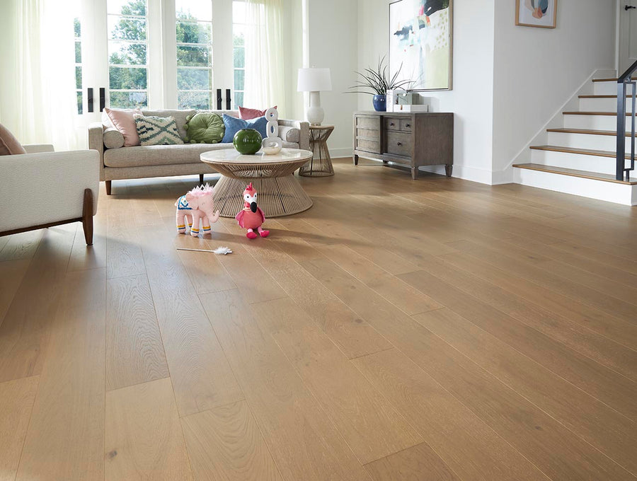 A spacious living room with light wood flooring, a beige sofa, armchair, round coffee table, and toys on the floor. Large windows with sheer curtains let in natural light. A staircase is visible on the right.