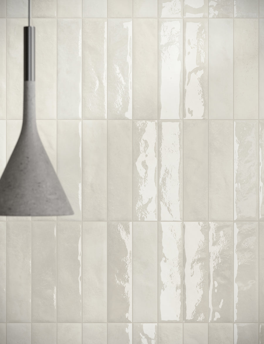 A modern pendant lamp hangs in front of a wall covered with glossy, rectangular cream-colored tiles, reflecting light and creating a subtle, elegant pattern.
