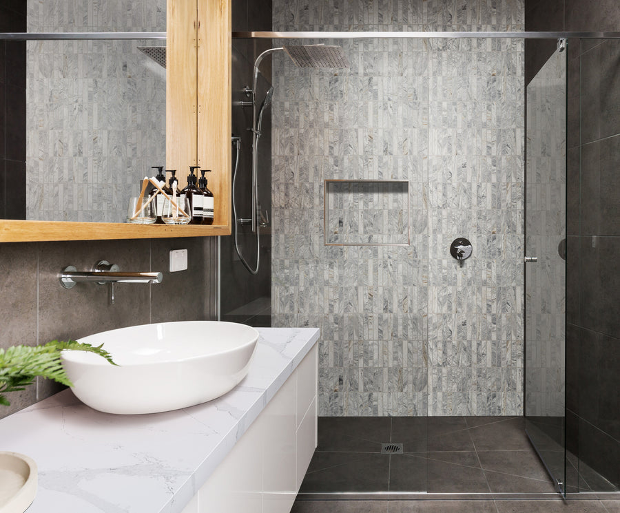 Modern bathroom with a white countertop and vessel sink, soap dispensers, and a plant. The walk-in shower has glass doors, grey textured tiles, a built-in niche, and a rainfall showerhead.