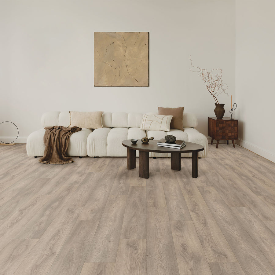 A modern living room with a white sofa, beige and brown throw pillows, a dark round coffee table, abstract wall art, and a wooden side table with a dried branch arrangement, all on light wood flooring.