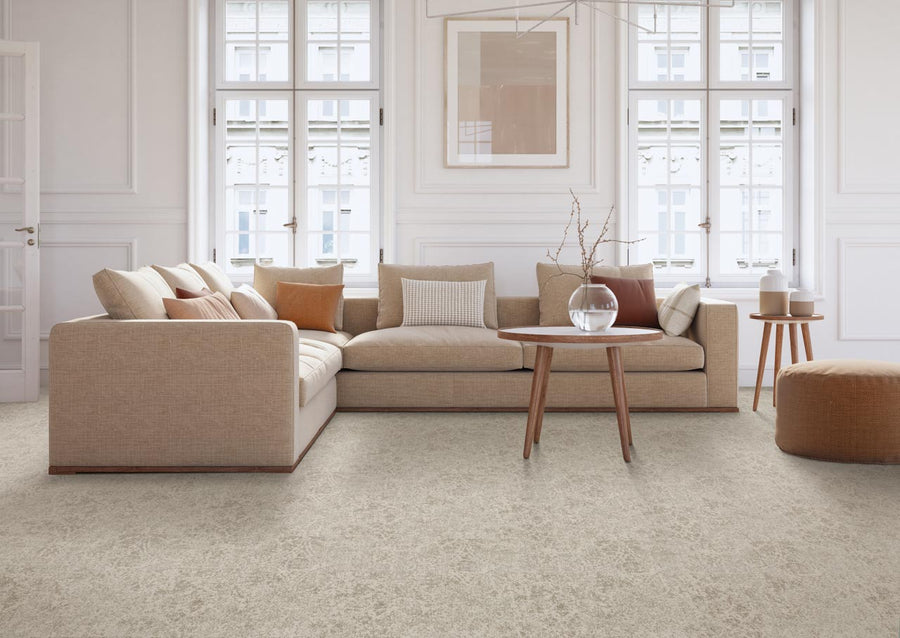 A modern living room features a beige sectional sofa, neutral and rust-colored pillows, a round wooden table with a glass vase, a brown pouf, and large windows illuminating the luxurious Subtle Grandeur On Trend Carpet.