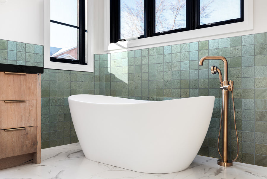 Modern bathroom with a white freestanding bathtub, green tiled walls, a copper floor-mounted faucet, wood cabinetry, and large windows letting in natural light. Marble-patterned floor tiles complete the space.