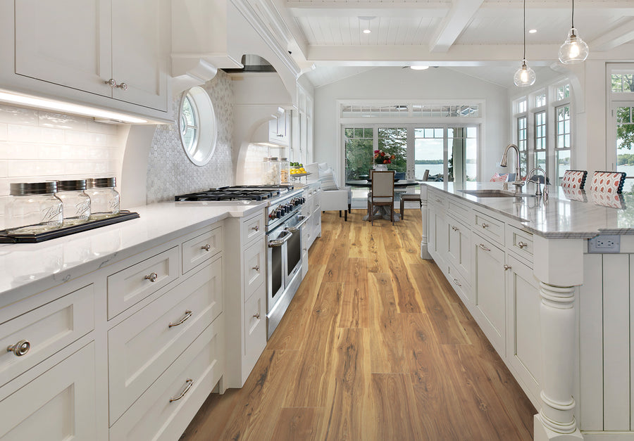 Bright, spacious kitchen with white cabinets, stainless steel appliances, a large island with barstools, wood flooring, pendant lights, and a view into a dining area with large windows overlooking greenery and water.