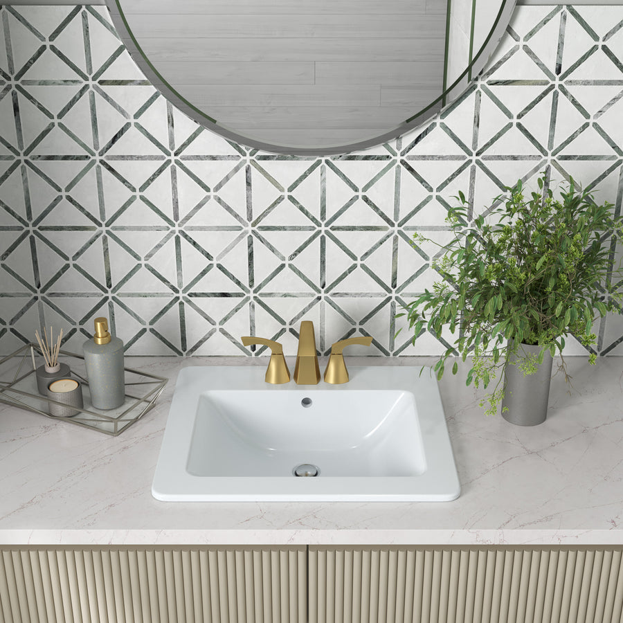 A modern bathroom sink with gold faucet, set in a white countertop. Behind it is a geometric patterned tile wall, a round mirror, a potted green plant, and a tray holding soap and reed diffusers.