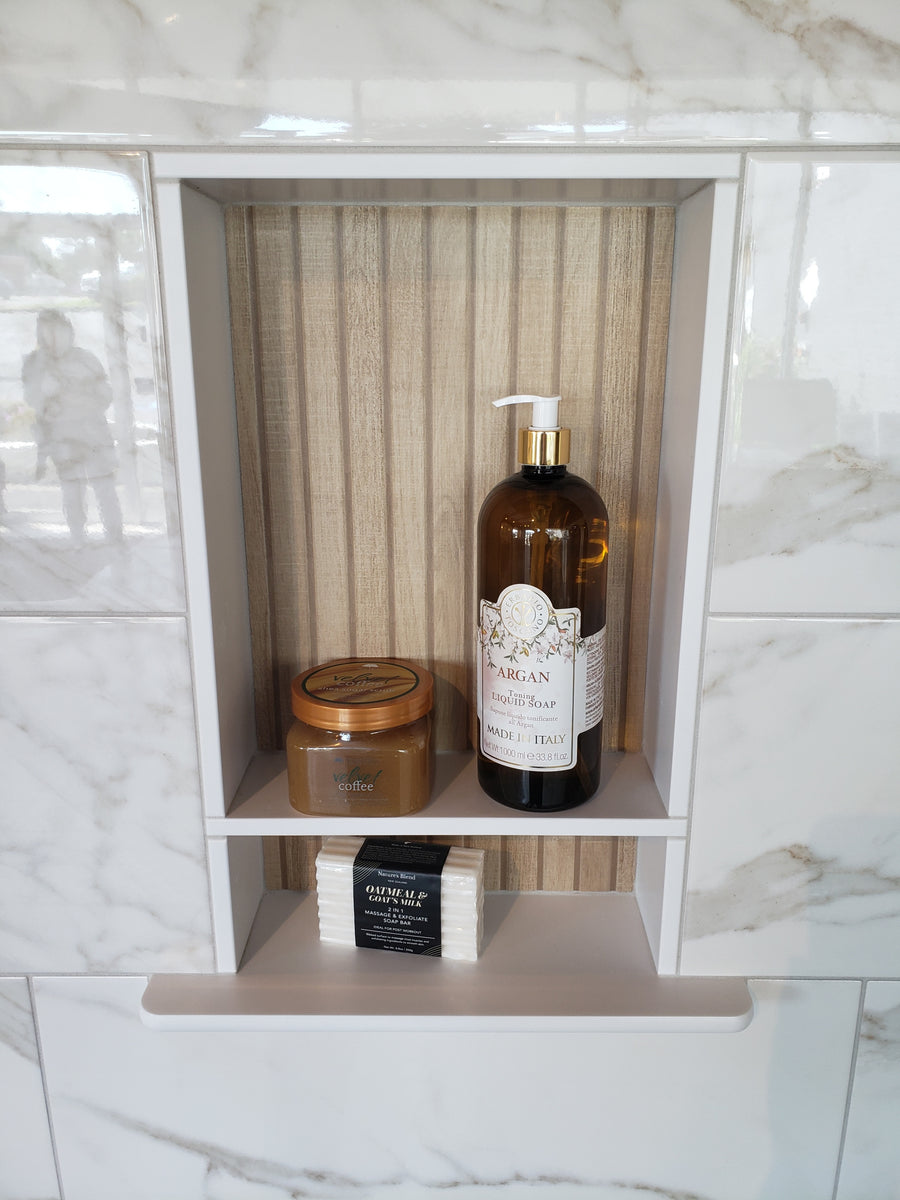 A shower niche with a marble tile surround contains a jar of body scrub, a large pump bottle of argan conditioner, and a white bar of soap on a shelf. The background is wood-patterned.