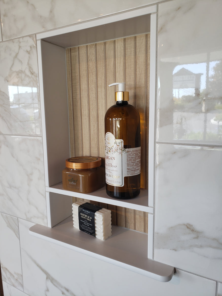 A shower niche with marble tile holds a large amber pump bottle, a small round jar, a black soap bar, and three stacked white soap bars on two shelves. A window reflection appears in the shiny tile.