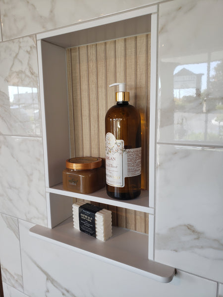 A shower niche with marble tile holds a large amber pump bottle, a small round jar, a black soap bar, and three stacked white soap bars on two shelves. A window reflection appears in the shiny tile.