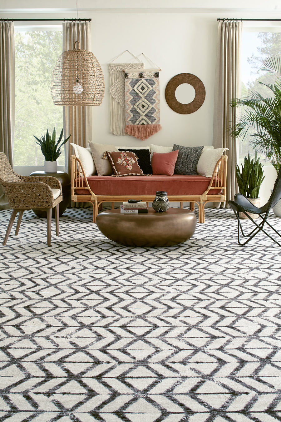 A stylish living room with a geometric-patterned rug, a wooden sofa with cushions, a round bronze coffee table, woven chair, indoor plants, and decorative wall art, lit by natural light from windows with beige curtains.