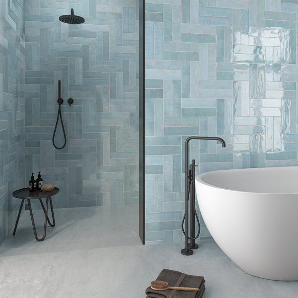 Modern bathroom with a freestanding white bathtub, matte black fixtures, and walk-in shower. Walls are covered in glossy light blue herringbone tiles; a small black stool and neatly placed towels complete the minimalist decor.
