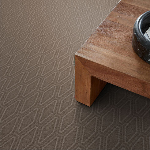 A close-up of a brown patterned carpet with a geometric diamond design, next to the corner of a wooden table that has a black decorative bowl on top.