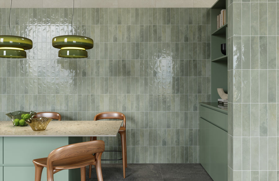 A modern kitchen with glossy green subway tiles on the walls, light green cabinets, wooden chairs, a beige countertop, and two green pendant lights. A bowl of green apples and a decorative glass bowl sit on the counter.
