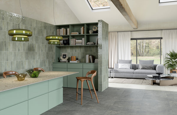Modern open-plan kitchen and living area with green cabinets, tiled walls, open shelves, two green pendant lights, and a wooden stool. Large windows and skylights let in natural light, and a gray sofa is in the background.